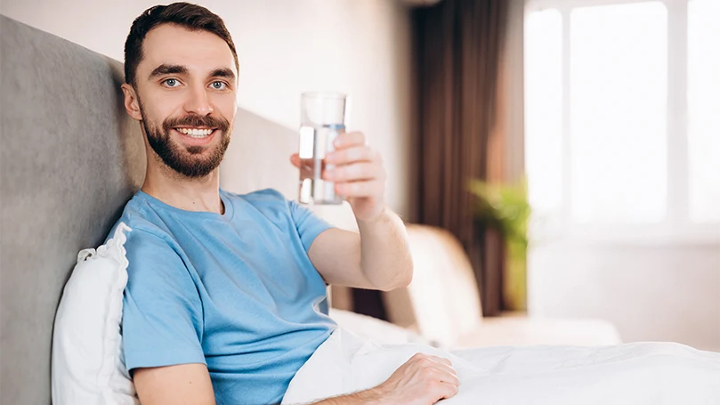 An image of a man drinking a glass of water in his bed before the bed time.