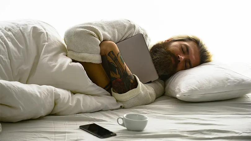 An image of a bearded man in deep sleep hugging tablet.