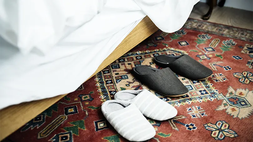 An image of a Persian rug next to a bed with two pairs of slippers on it