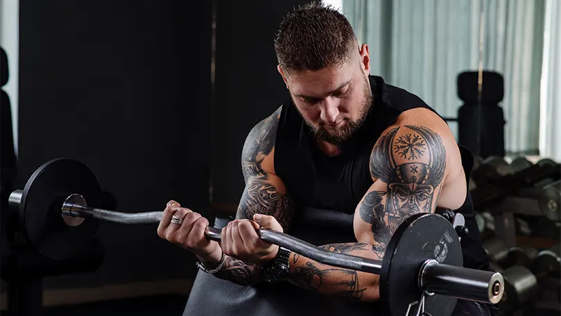 An image of a muscular man with tatooes lifting weights.