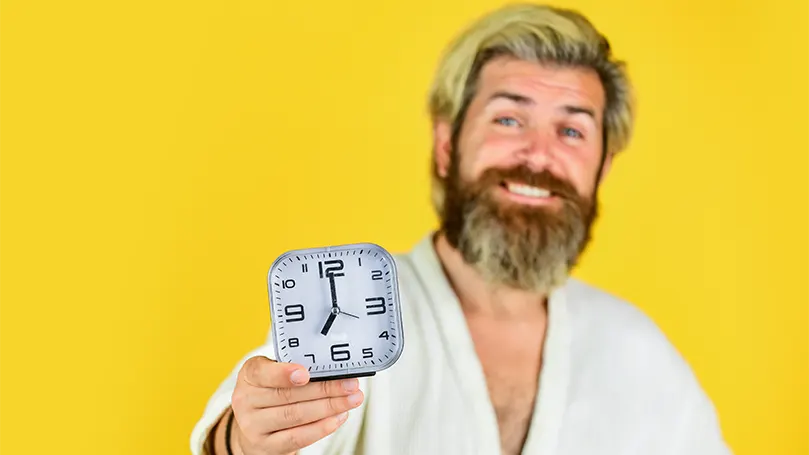 An image of a sleepy middle aged man with beard in a bathrobe holding an alarm clock.