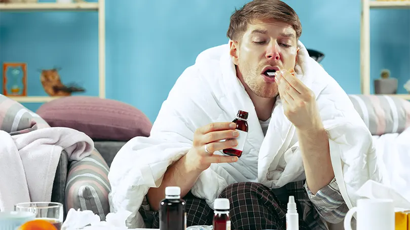 An image of a man taking cough syrup while covered with a blanket