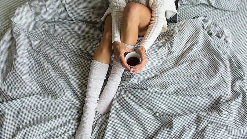An image of a woman in socks holding a cup of coffee in bed.