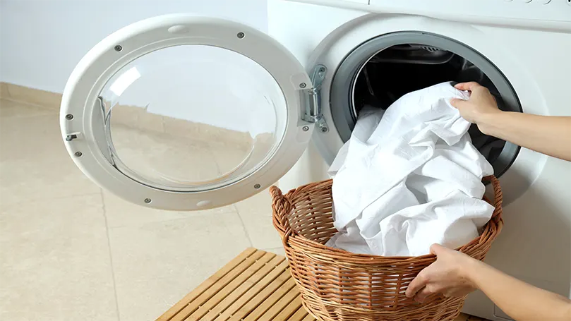 a woman putting white sheets in a washing machine.