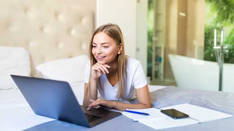 An image of a woman lying on her bed working on her laptop