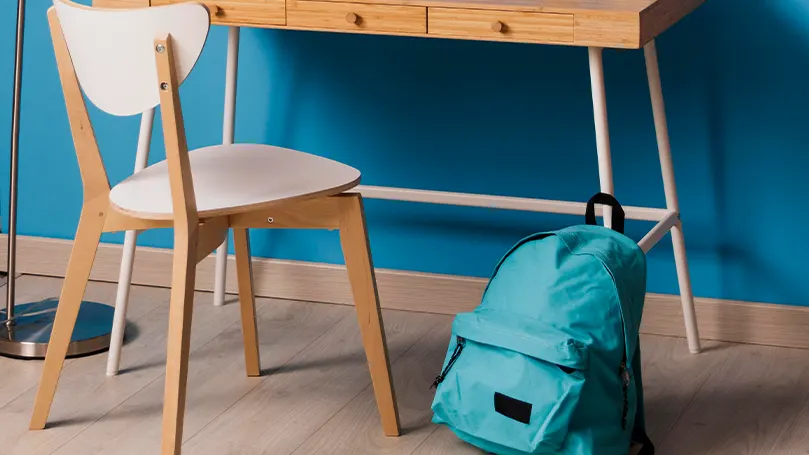 Blue wall, wooden floor with a wooden desk and chair. A blue rucksack placed in front.