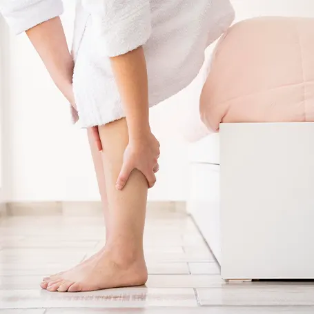 An image of a woman sitting on her bed and holding her leg in pain