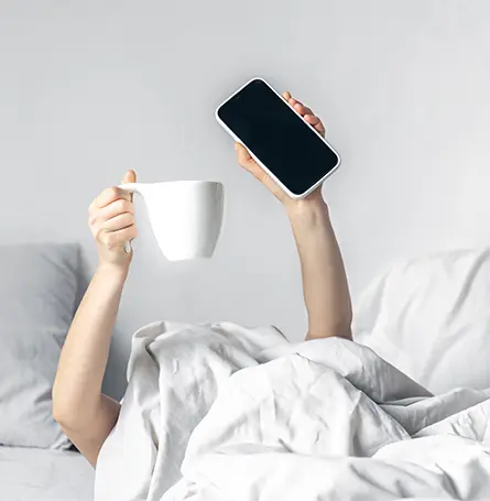 An image of a person in bed holding up a mug and their phone