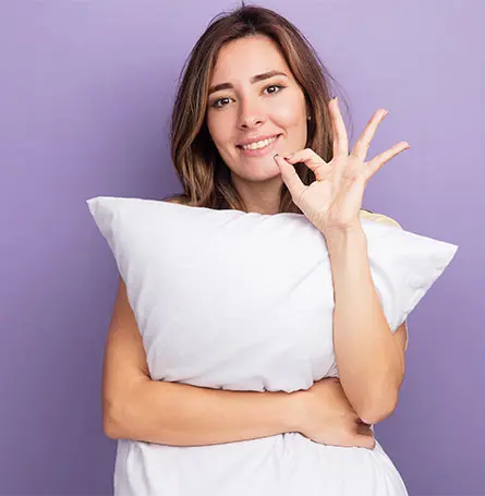 An image of a woman holding a pillow and holding up the okay sign